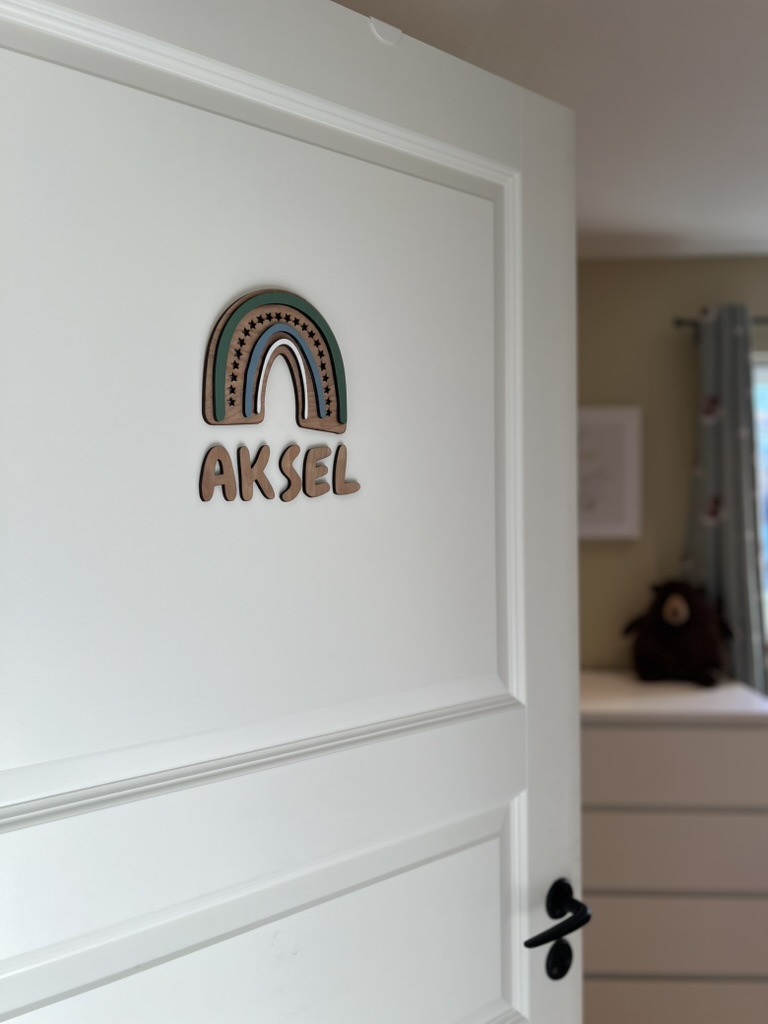 Navneskilt til barnerom med en håndlaget blå regnbue som henger over barnets navn.
