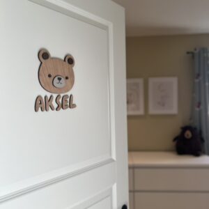 Et navneskilt på barnerommet med personlig, håndlaget navneskilt med bamse og navnet Aksel på hvit dør.