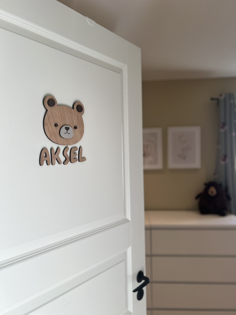 Et navneskilt på barnerommet med personlig, håndlaget navneskilt med bamse og navnet Aksel på hvit dør.