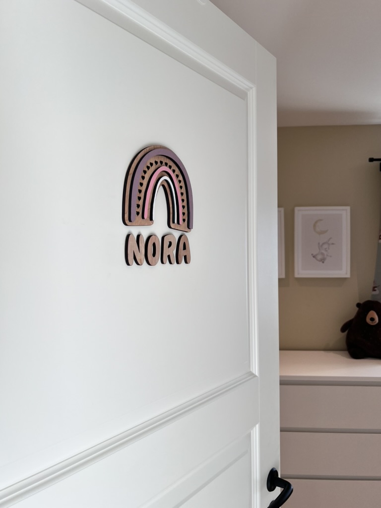 Navneskilt til barnerom med en håndlaget rosa regnbue som henger over barnets navn.