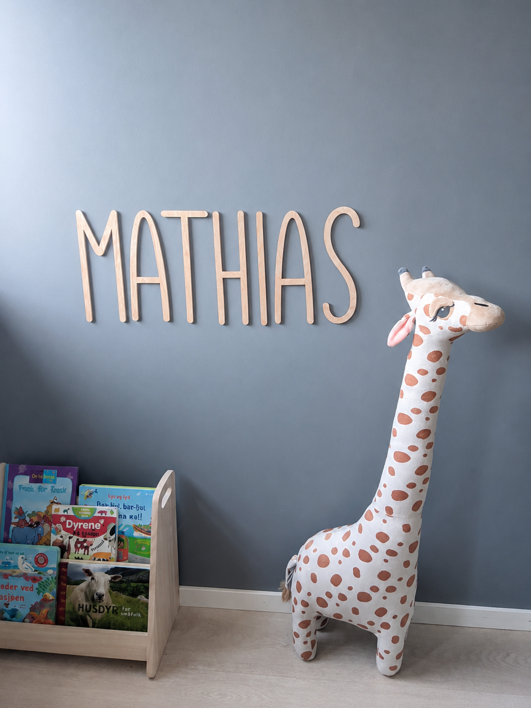 Barnerom med navnet «MATHIAS» i trebokstaver på blågrå vegg, en stor giraffkrammedyr og en bokhylle med barnebøker.