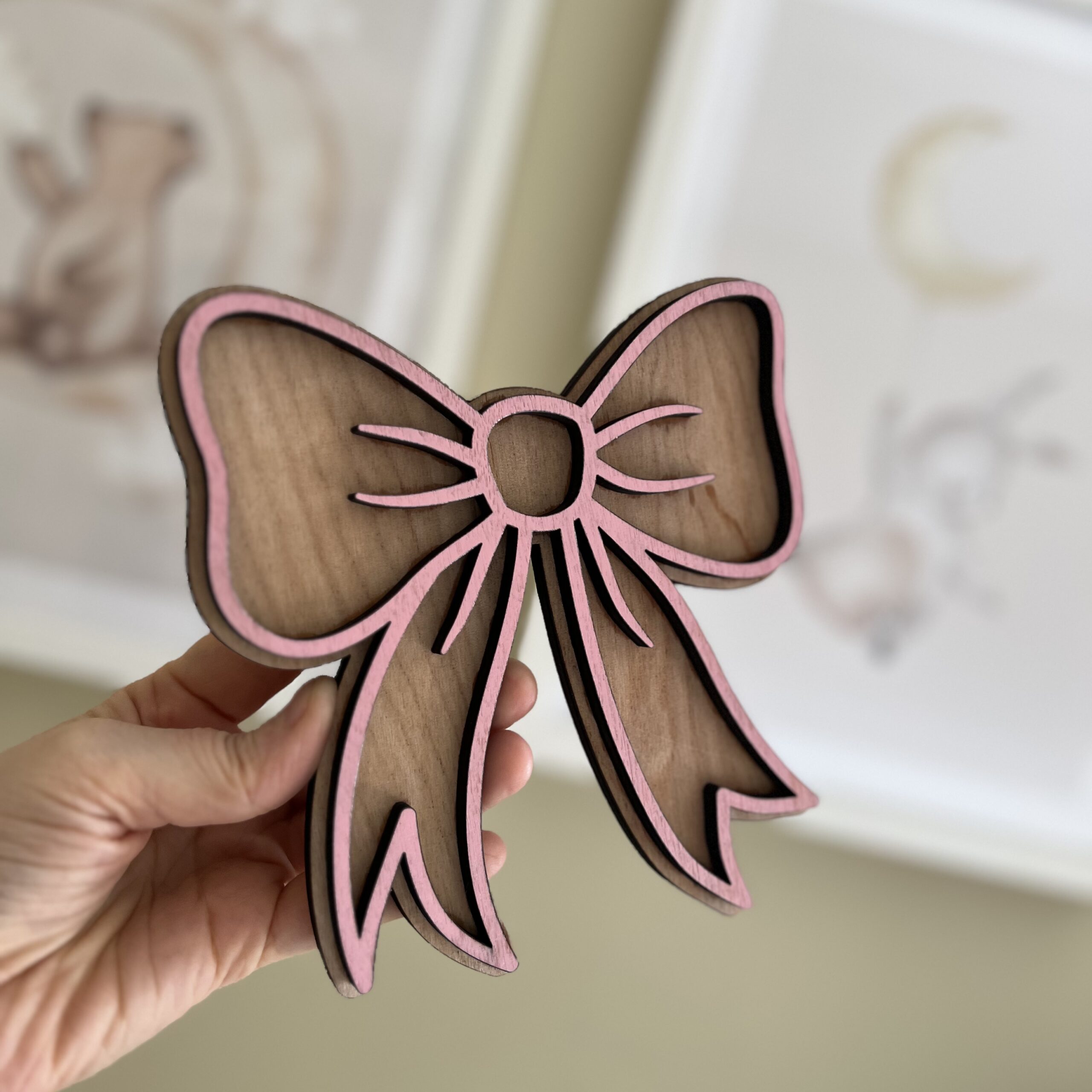 Håndlaget sløyfeformet veggdekor fra ByRossland i bjørkfiner med rosa detaljer, perfekt som personlig pynt på barnerommet.