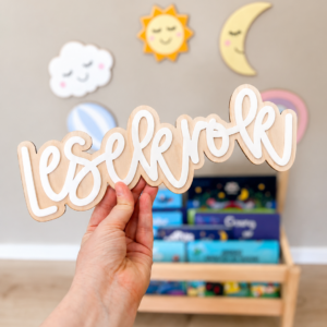 En hånd holder et treskiltstilt med teksten «lesekrok» foran en barnebok-hylle og veggdekorasjoner med sol, måne og sky.