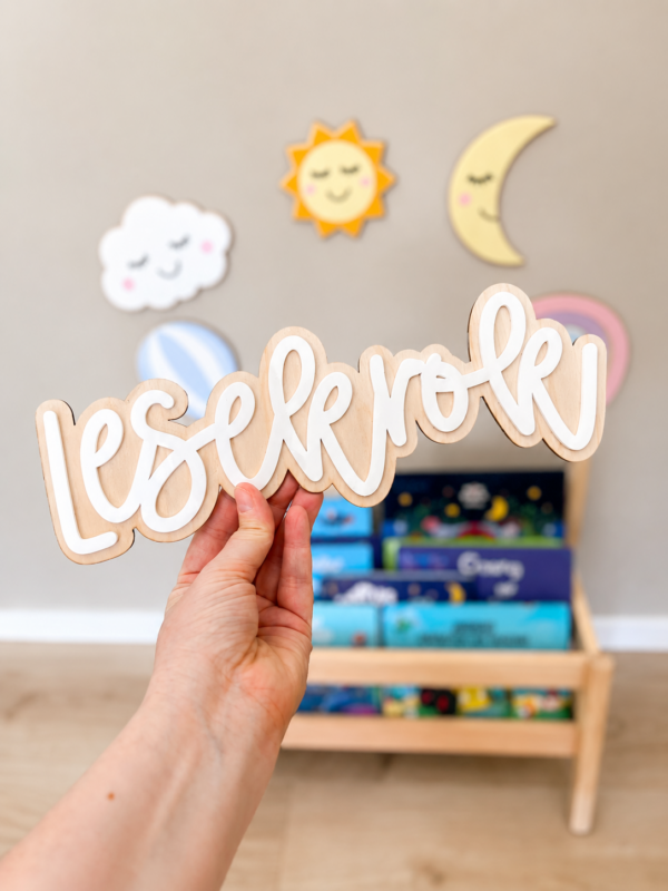 En hånd holder et treskiltstilt med teksten «lesekrok» foran en barnebok-hylle og veggdekorasjoner med sol, måne og sky.