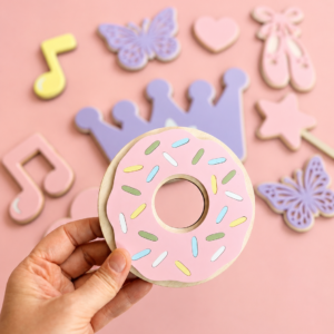 En hånd som holder opp en donut med rosa glasur. I bakgrunnen ser man annen veggdekor i pastellfarger på en rosa bakgrunn.