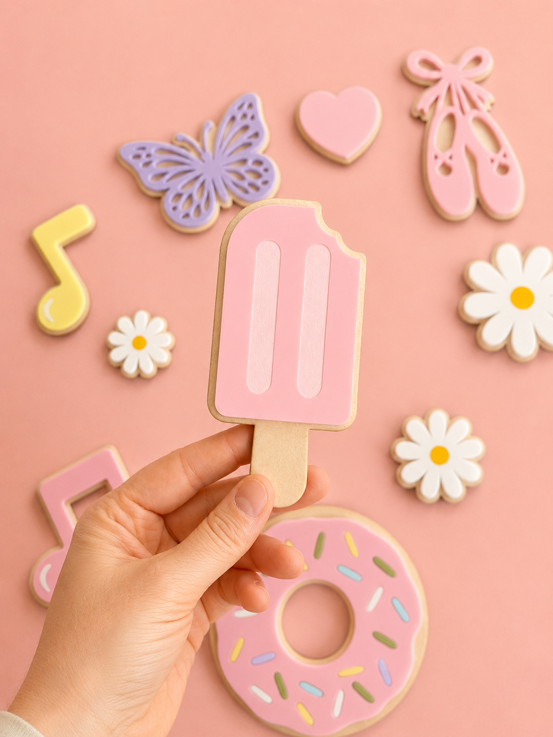 En hånd holder en rosa saftis, omgitt av fargerik veggdekor i form av sommerfugl, blomster, donut, balletsko og musikknote.