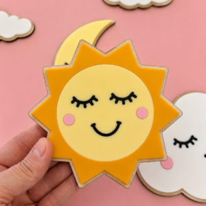 En hånd holder en solformet veggdekor med smilende ansikt, omgitt av sky- og måne på rosa bakgrunn.