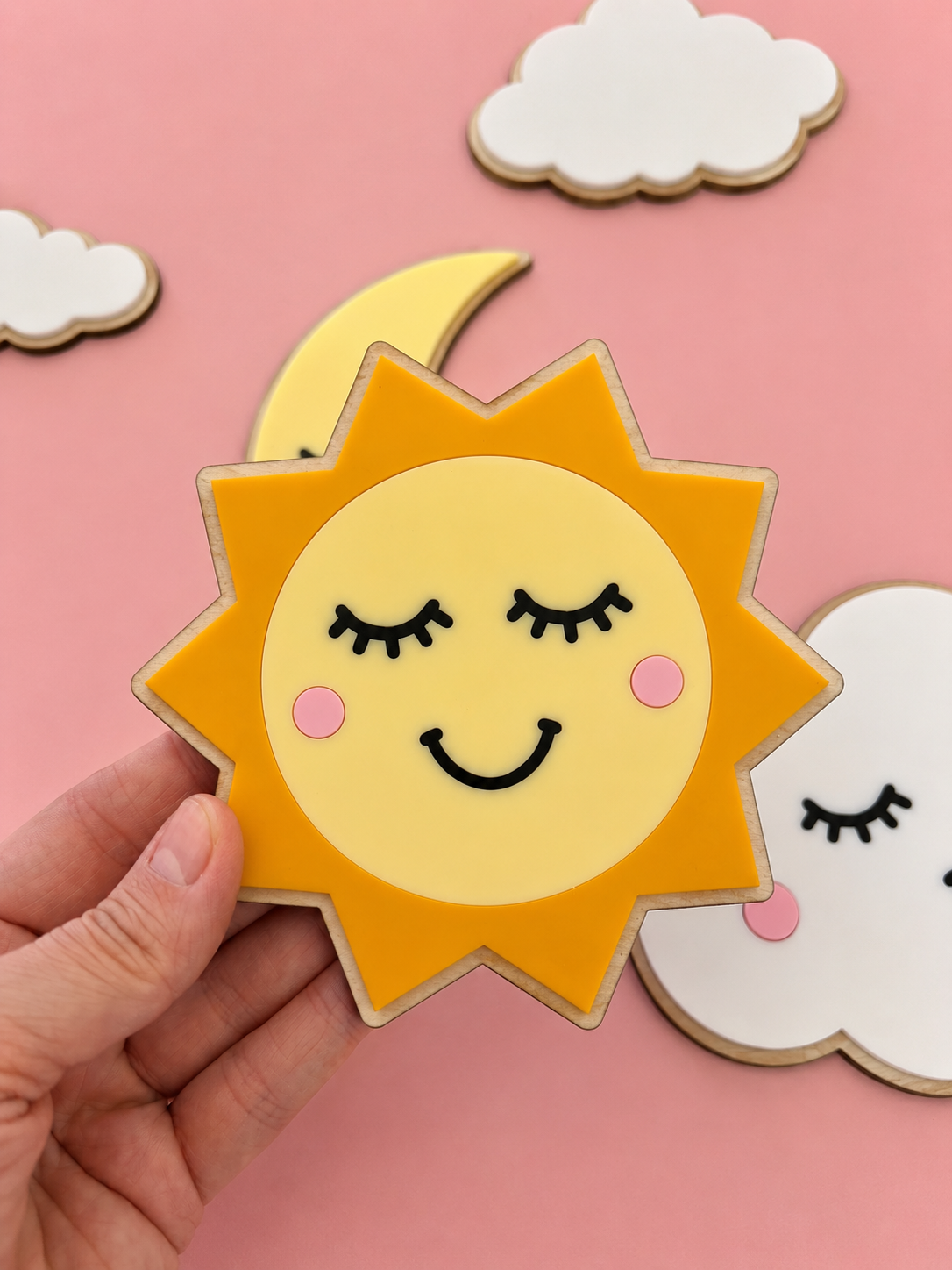En hånd holder en solformet veggdekor med smilende ansikt, omgitt av sky- og måne på rosa bakgrunn.
